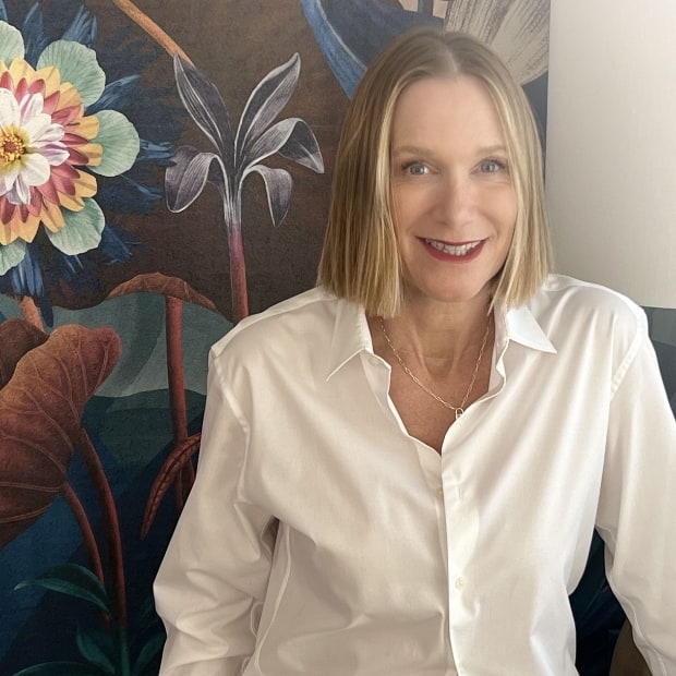 Carolyn-mural-real-620x620-1 alt= A woman with shoulder-length blonde hair, wearing a white shirt, smiles while sitting in front of a dark floral background designed by Byrdview-Webdesign.